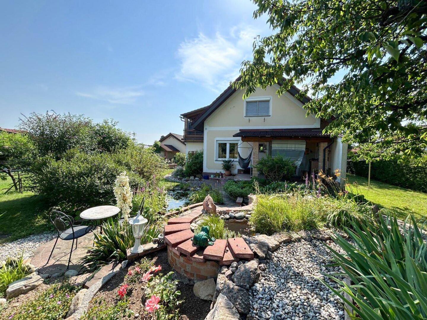 Weitläufiger Gartenbereich mit Zierteich, Sitzgelegenheiten und blühenden Pflanzen vor dem Haus.
