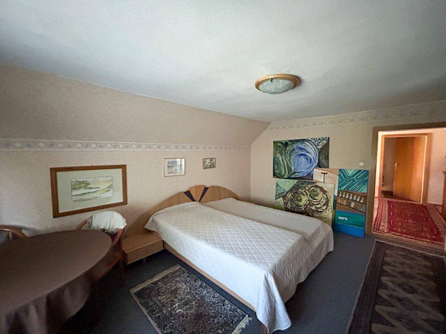 Gemütliches Schlafzimmer mit Doppelbett, Tisch und Dachschräge, dekoriert mit Kunstwerken.
