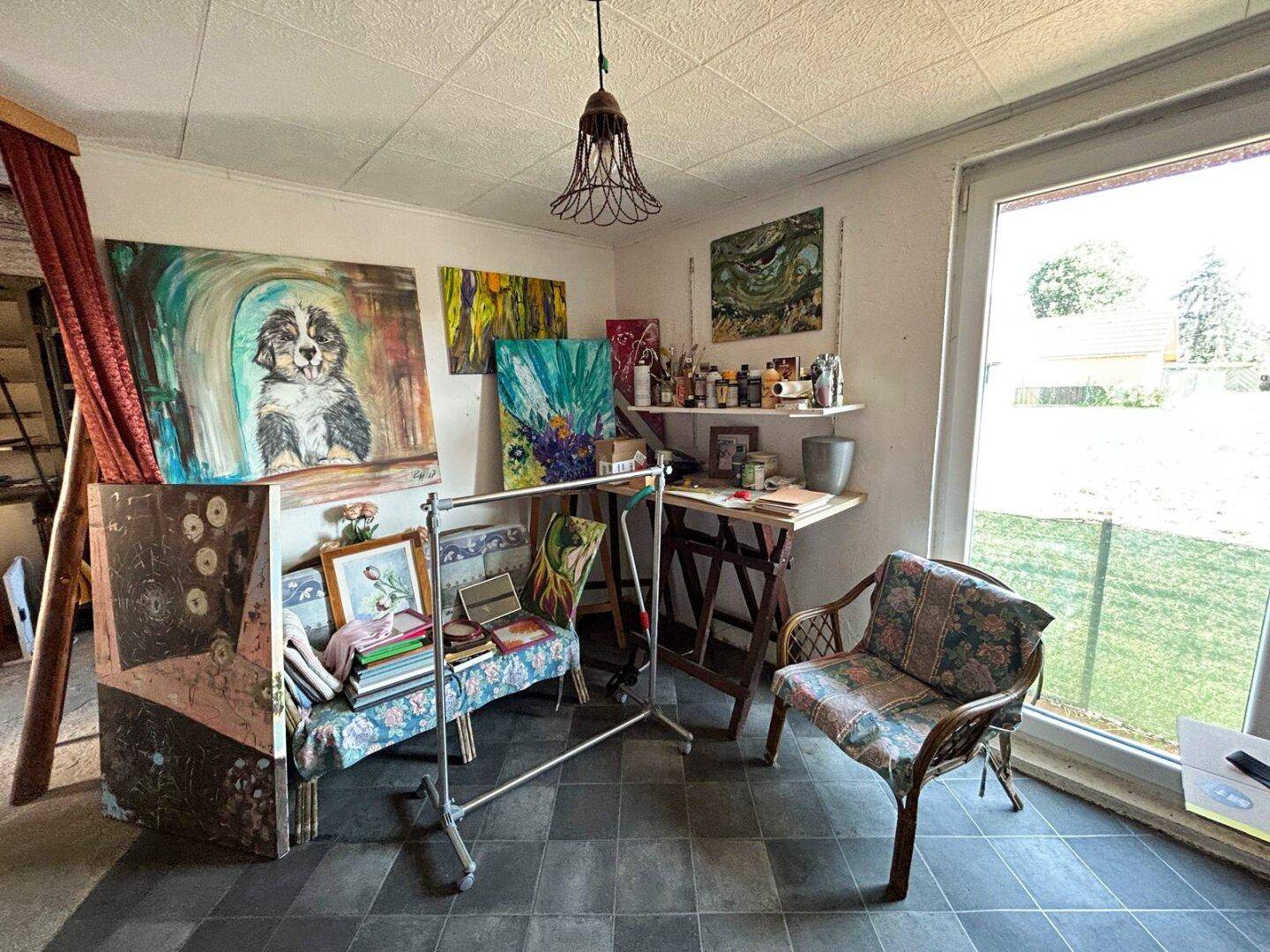 Künstleratelier mit verschiedenen Gemälden, Arbeitsbereich und großem Fenster.