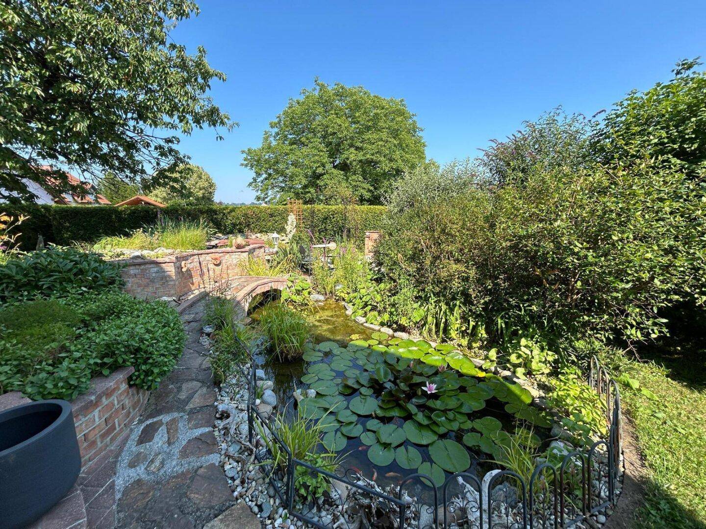 Gepflegter Garten mit Teich, Ziegelbrücke und vielfältiger Bepflanzung unter blauem Himmel.