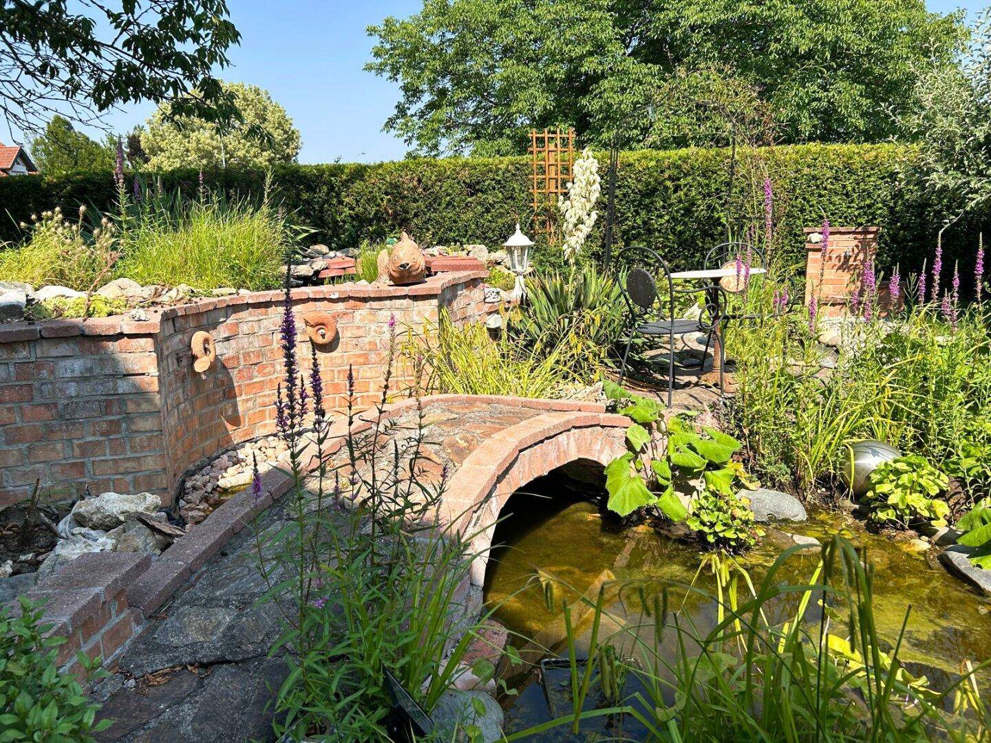 Detailansicht des Gartenteichs mit kleiner Ziegelbrücke und üppiger Bepflanzung.