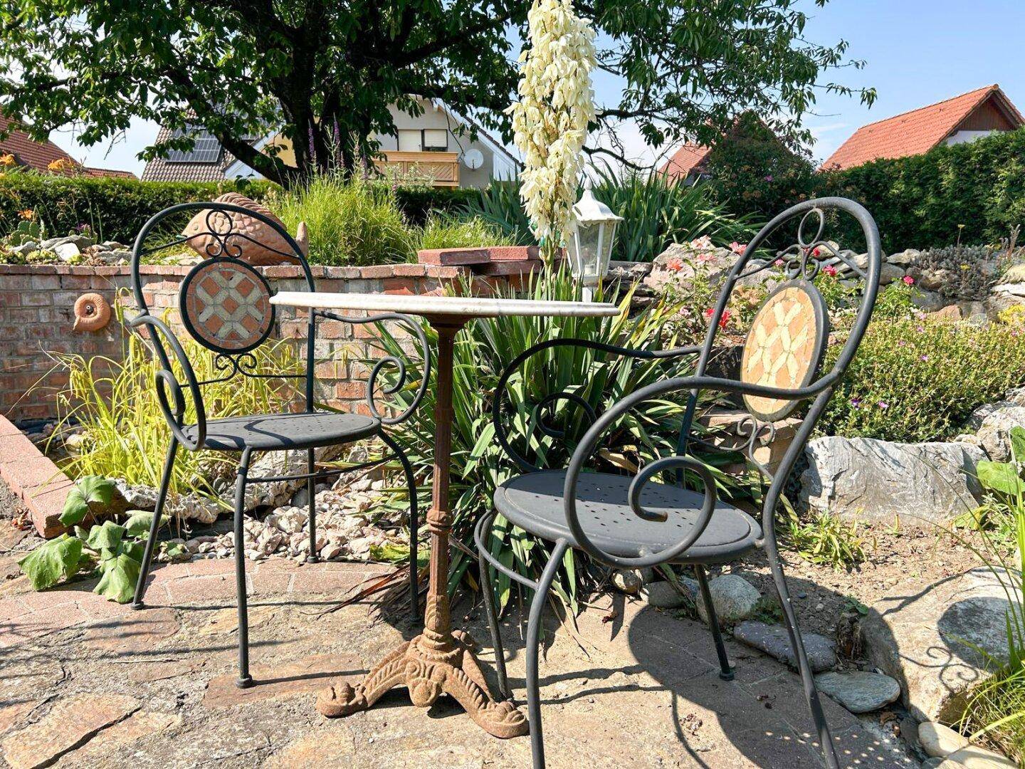 Gemütliche Sitzecke im Garten mit schmiedeeisernen Möbeln und Blumen.