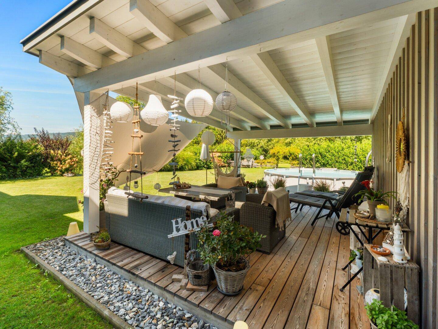Große überdachte Holzterrasse mit Lounge-Möbeln und Blick auf den Pool und Garten.