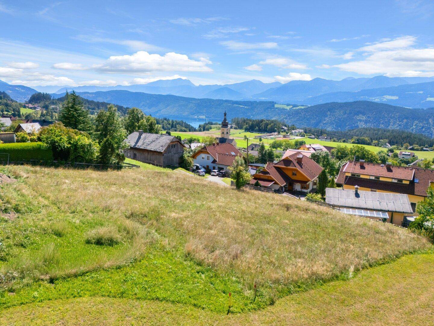 Weitläufige grüne Landschaft mit Häusern, einem Kirchturm und einem See im Hintergrund.