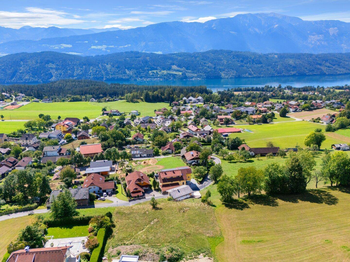 Weitläufige Luftaufnahme einer Siedlung am Seeufer, umgeben von grünen Hügeln und majestätischen Bergen.