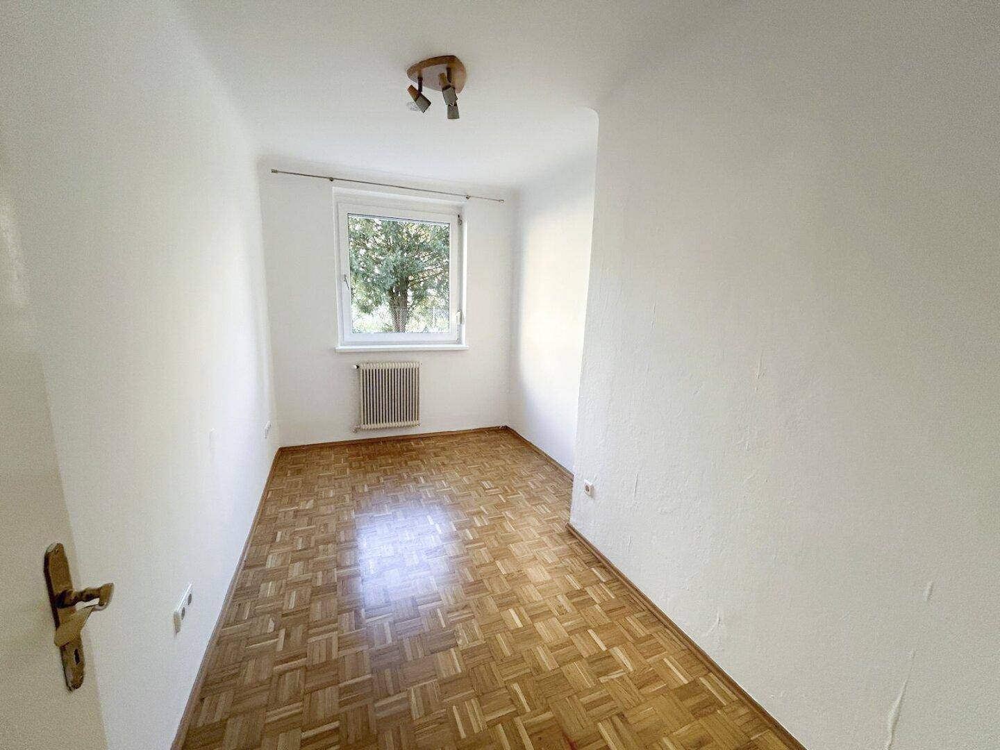Kleines, helles Zimmer mit Parkettboden und Fenster mit Blick auf Bäume.