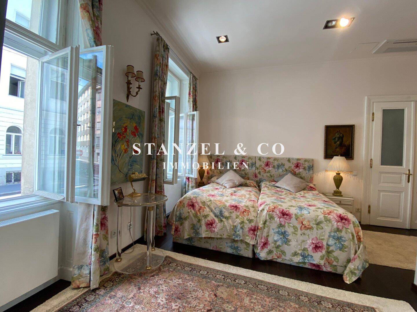 Helles Schlafzimmer mit floralen Mustern, Parkettboden und großen Fenstern.