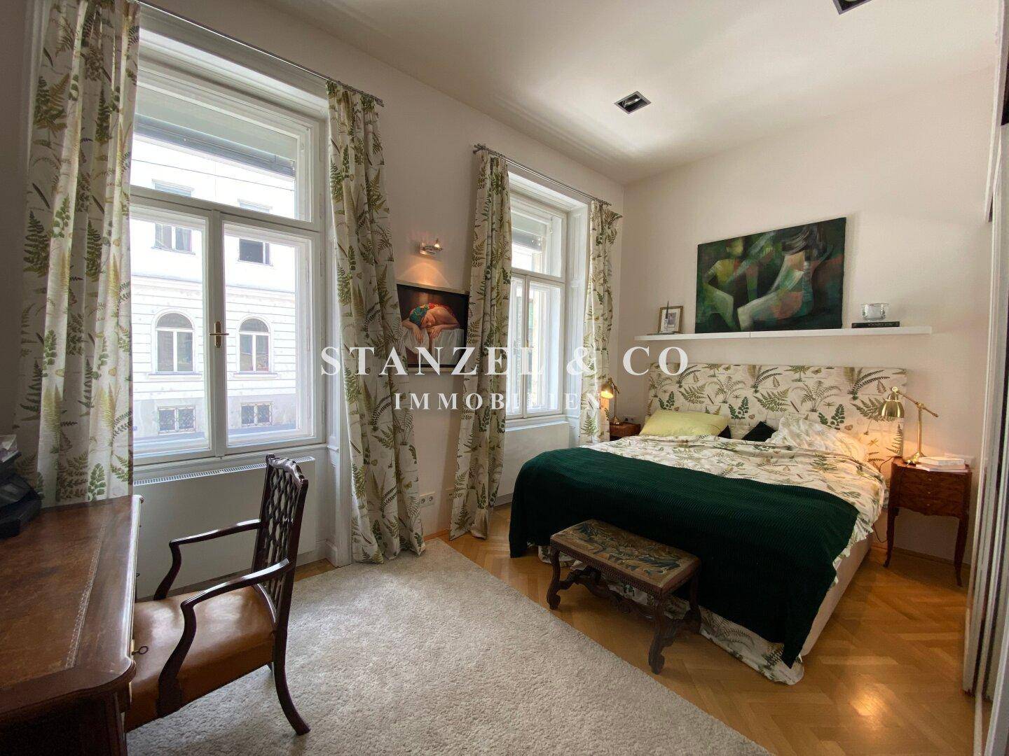 Helles Schlafzimmer mit Parkettboden, großen Fenstern und floralen Vorhängen.