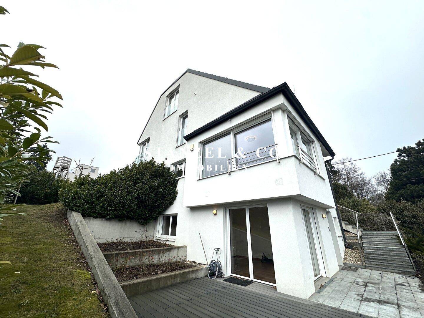 Modernes Haus mit weißer Fassade, großer Holzterrasse und Zugang zum Gartenbereich.