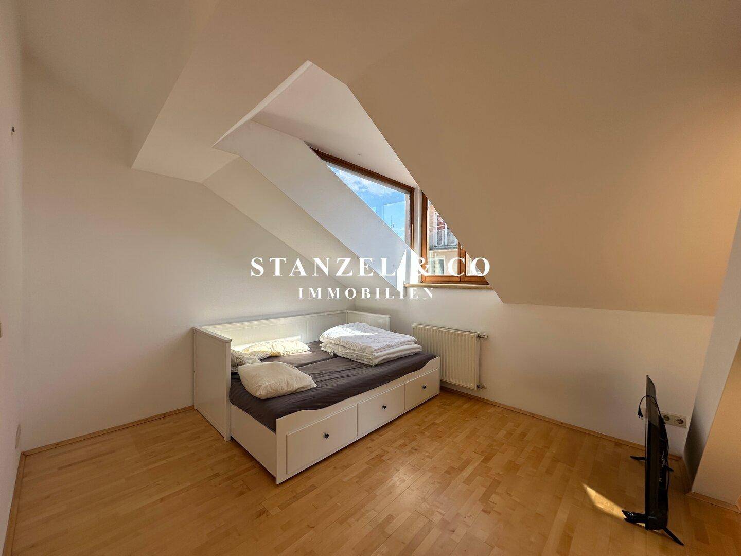 Schlafbereich im Dachgeschoss mit Holzboden, Bett und einem Dachfenster, das den Raum erhellt.
