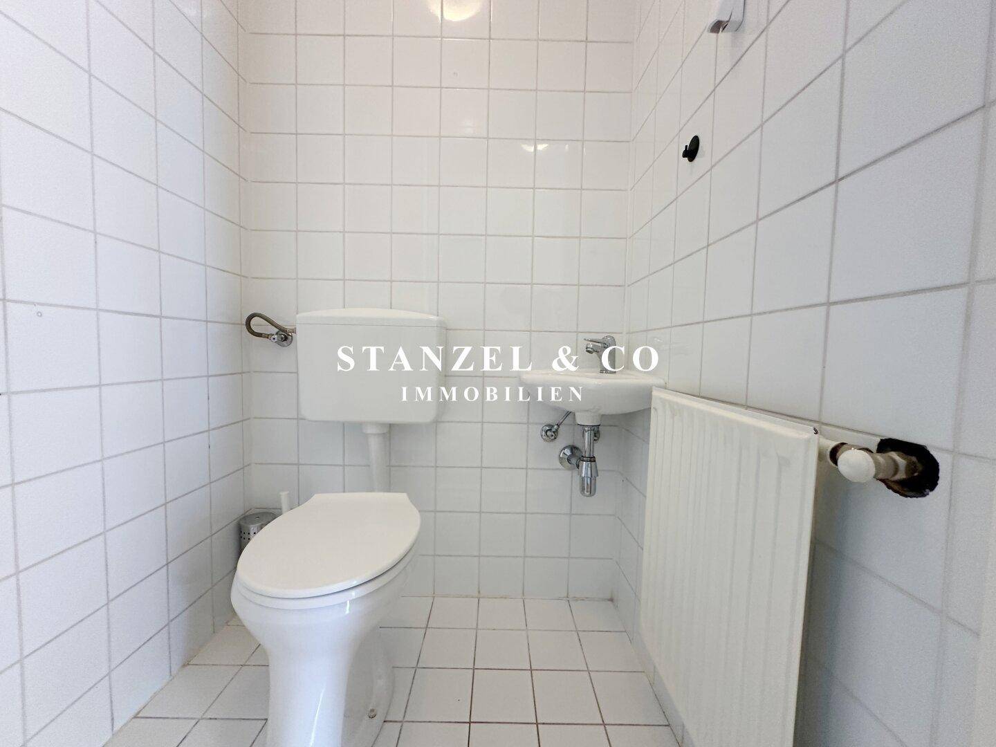 Separates WC mit weißen Fliesen, Toilette und kleinem Handwaschbecken.