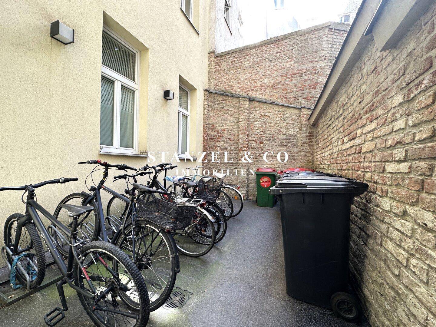 Außenbereich mit Fahrradabstellplätzen und Mülltonnen vor einer Ziegelwand.