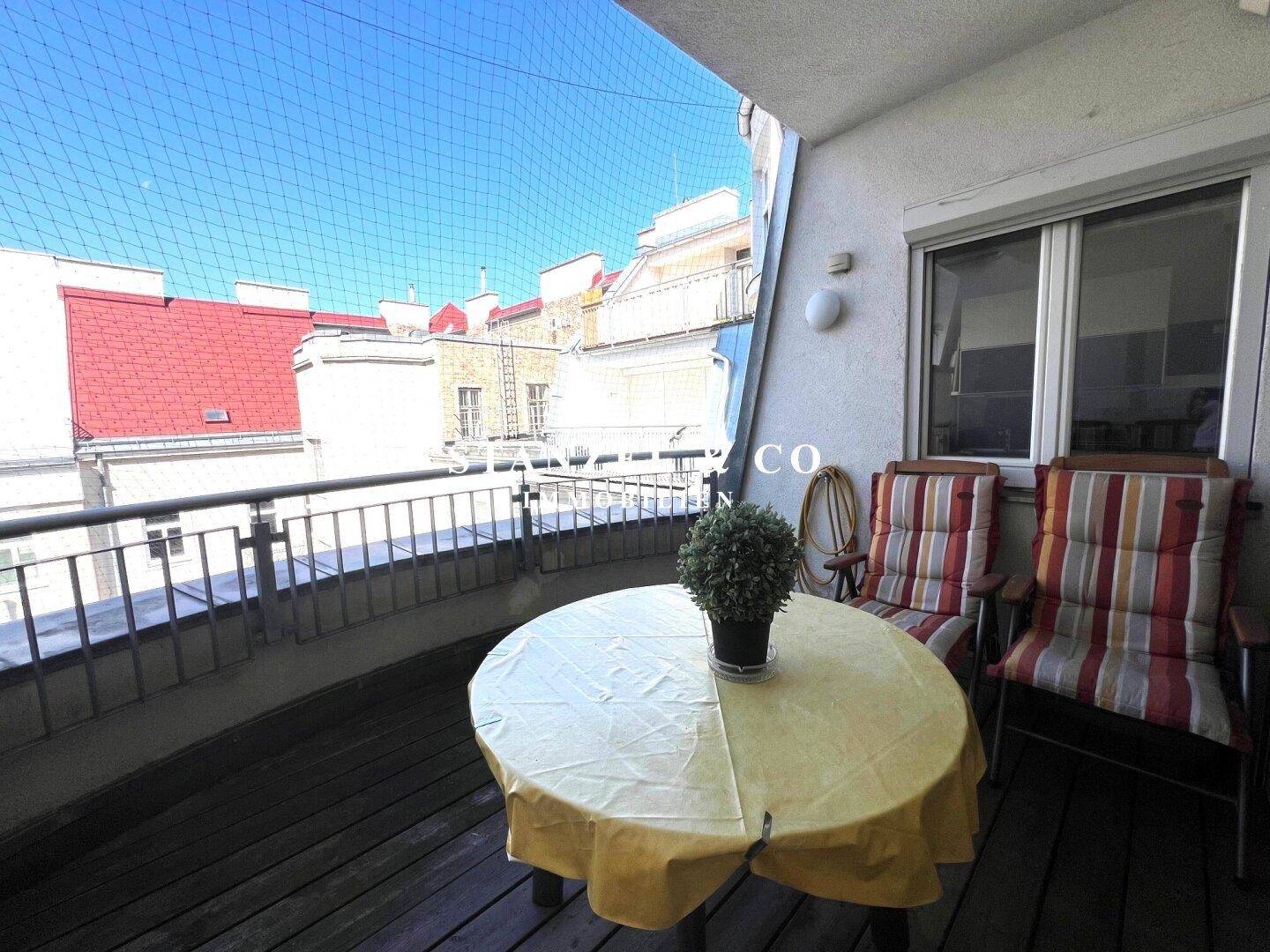 Heller Balkon mit Holzboden und freiem Blick auf den blauen Himmel über den Dächern.