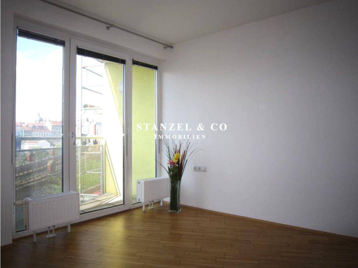 Helles, leeres Zimmer mit Parkettboden, weißen Wänden und Zugang zum Balkon.