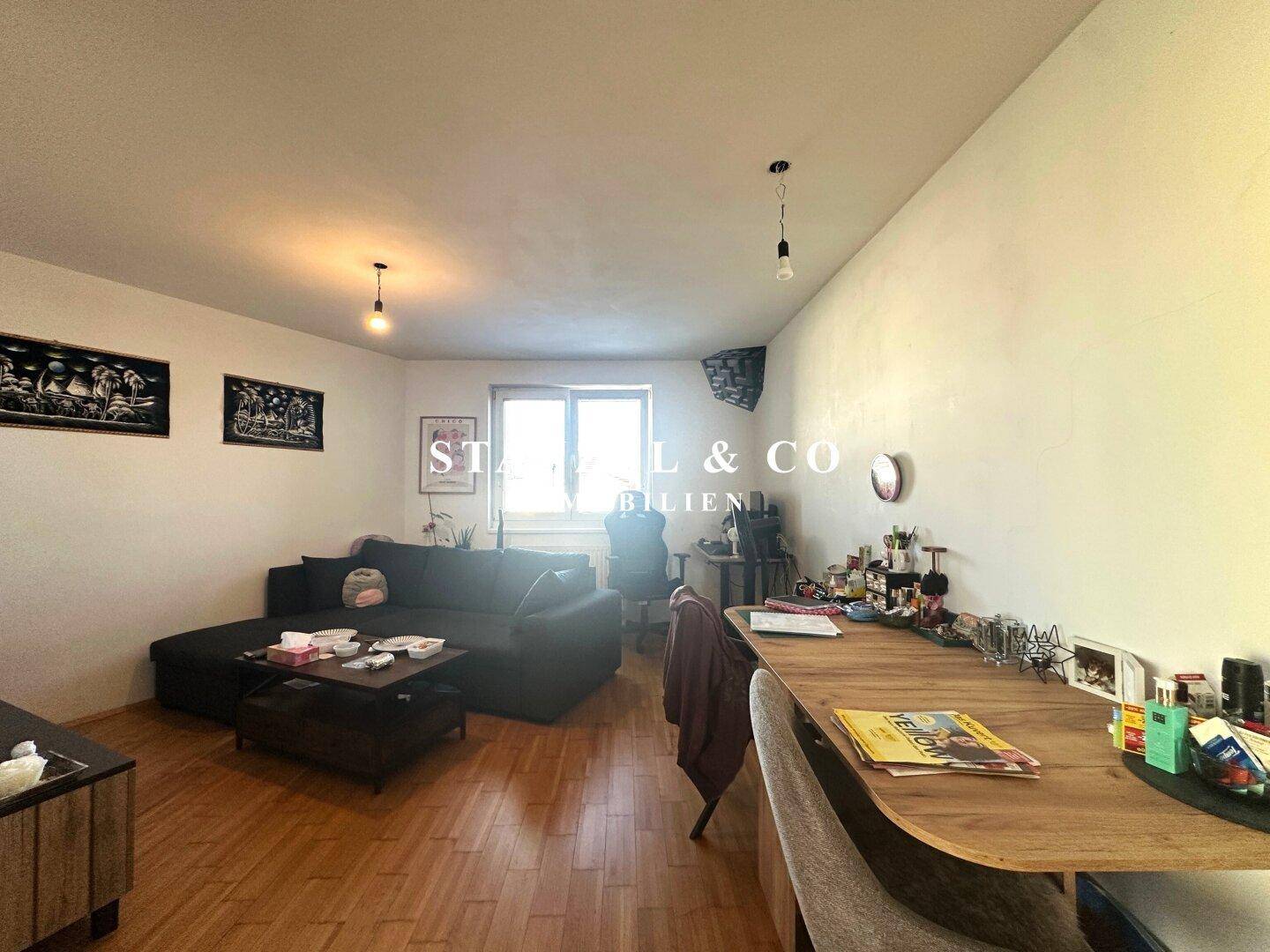 Gemütliches Wohnzimmer mit dunklem Sofa, Holzboden und einem Arbeitsbereich am Fenster.