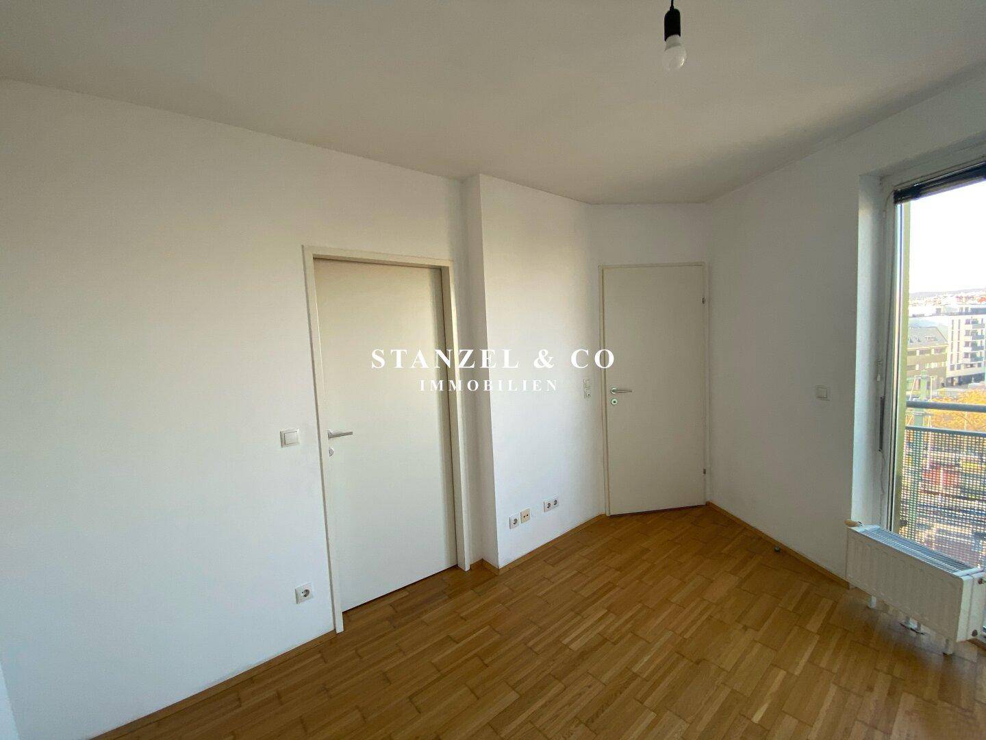Leeres Zimmer mit Parkettboden, weißen Wänden und Zugang zu einem Balkon oder einer Terrasse.
