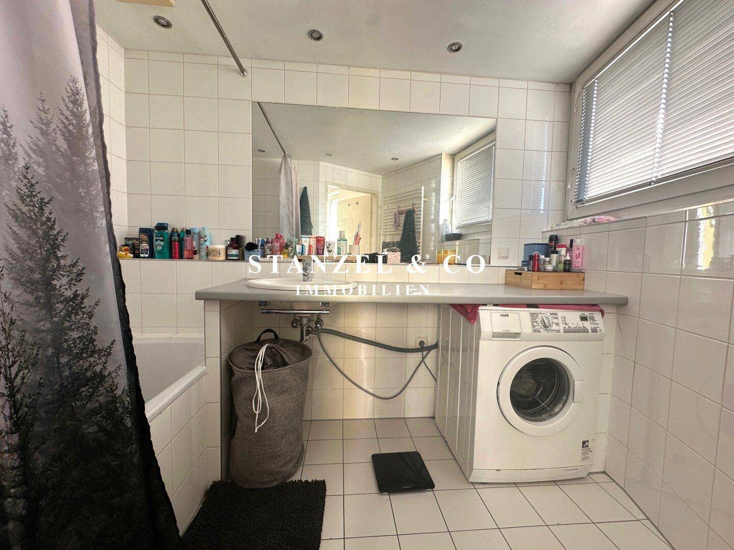 Funktionales Badezimmer mit Badewanne, Waschbecken und Waschmaschine, gefliest in Weiß und Grau.