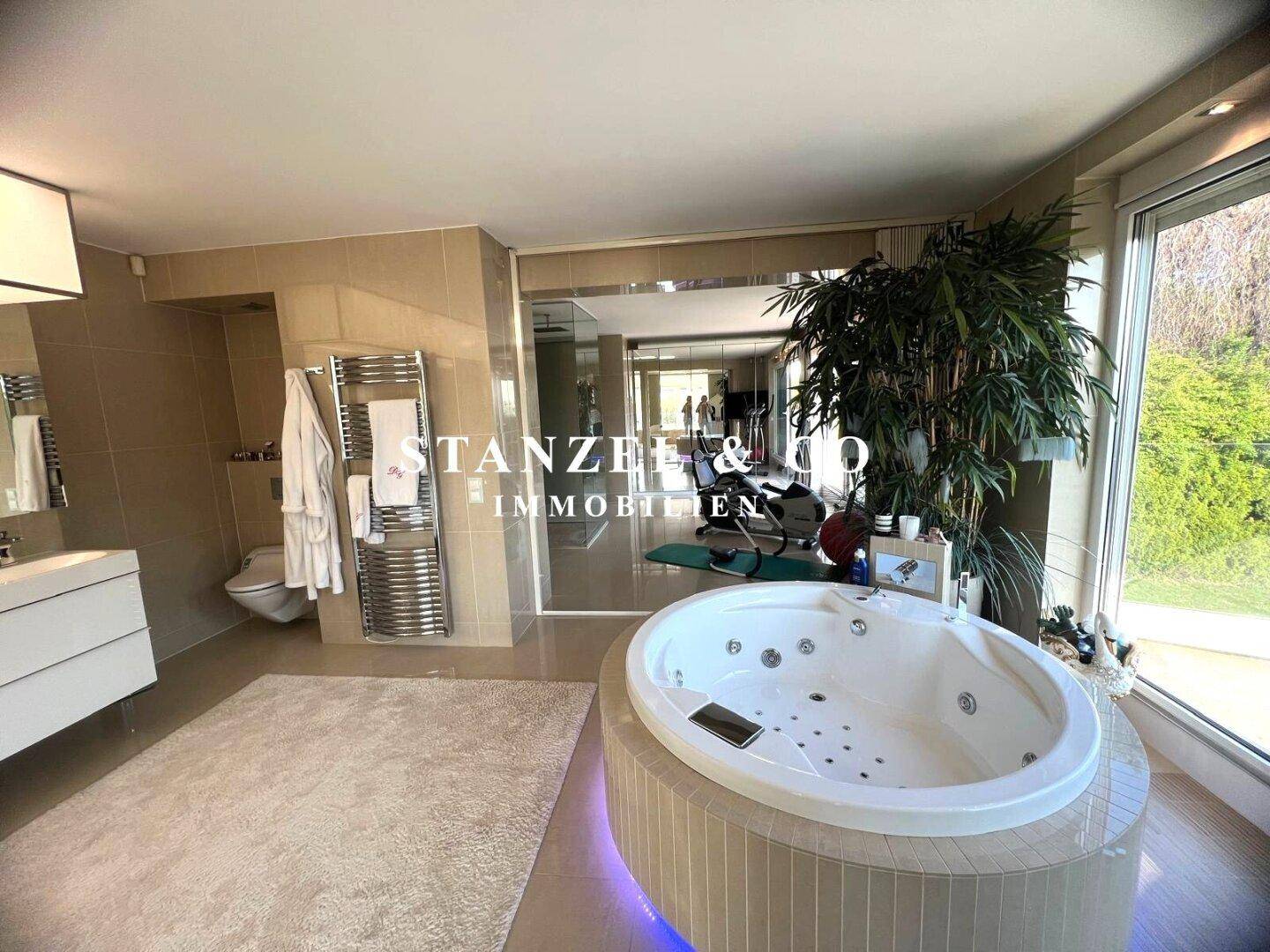 Luxuriöses Badezimmer mit großem Whirlpool, Doppelwaschbecken und raumhohen Fenstern mit Grünblick.