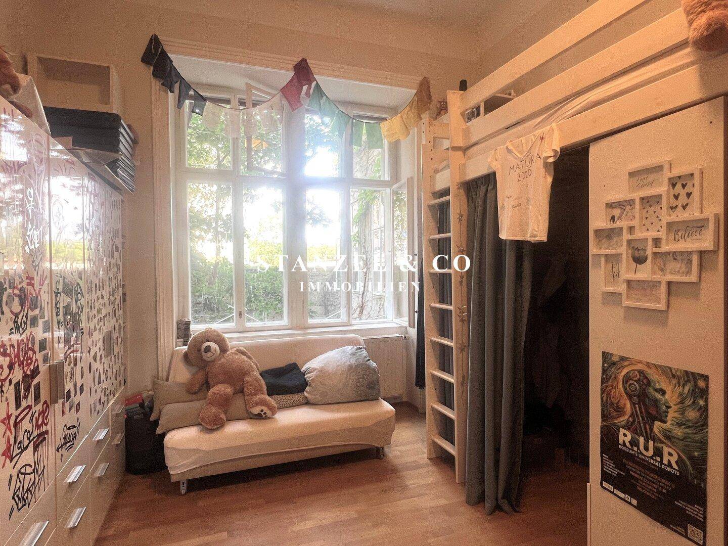 Helles Schlafzimmer mit Hochbett, Sofa und großem Fenster mit Blick ins Grüne.