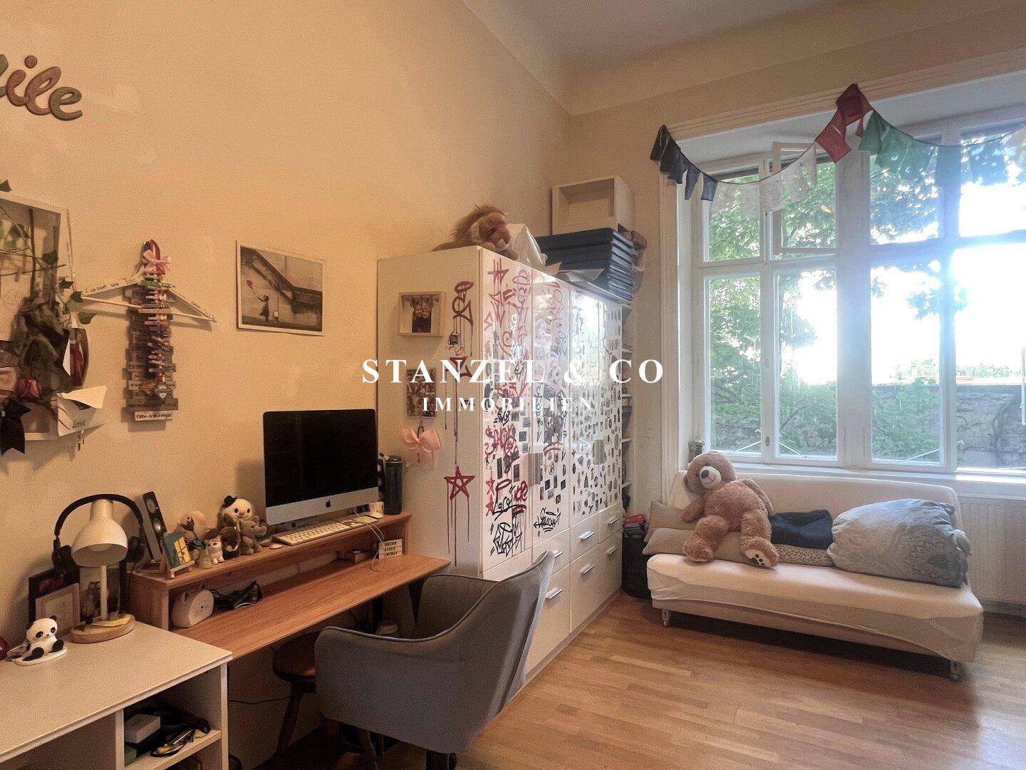 Helles Zimmer mit großem Fenster, Schreibtisch, Kleiderschrank und kleiner Couch, ideal als Kinderzimmer.