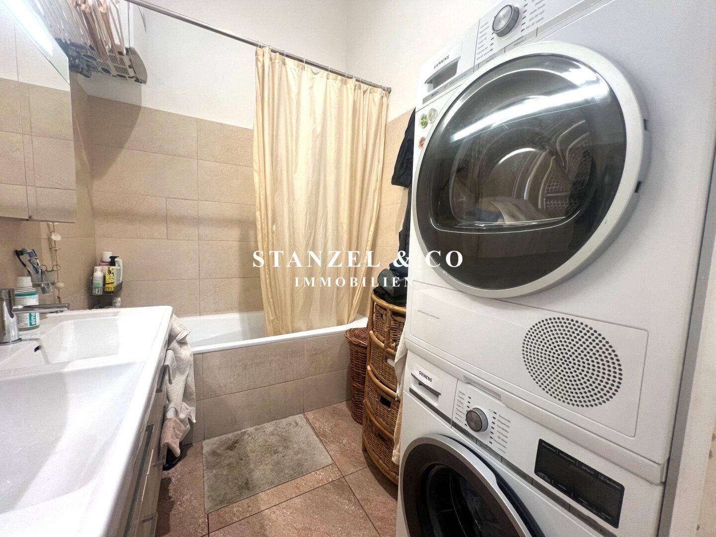 Badezimmer mit Badewanne, Waschbecken und praktischer Waschmaschine sowie Trockner, hell gefliest.