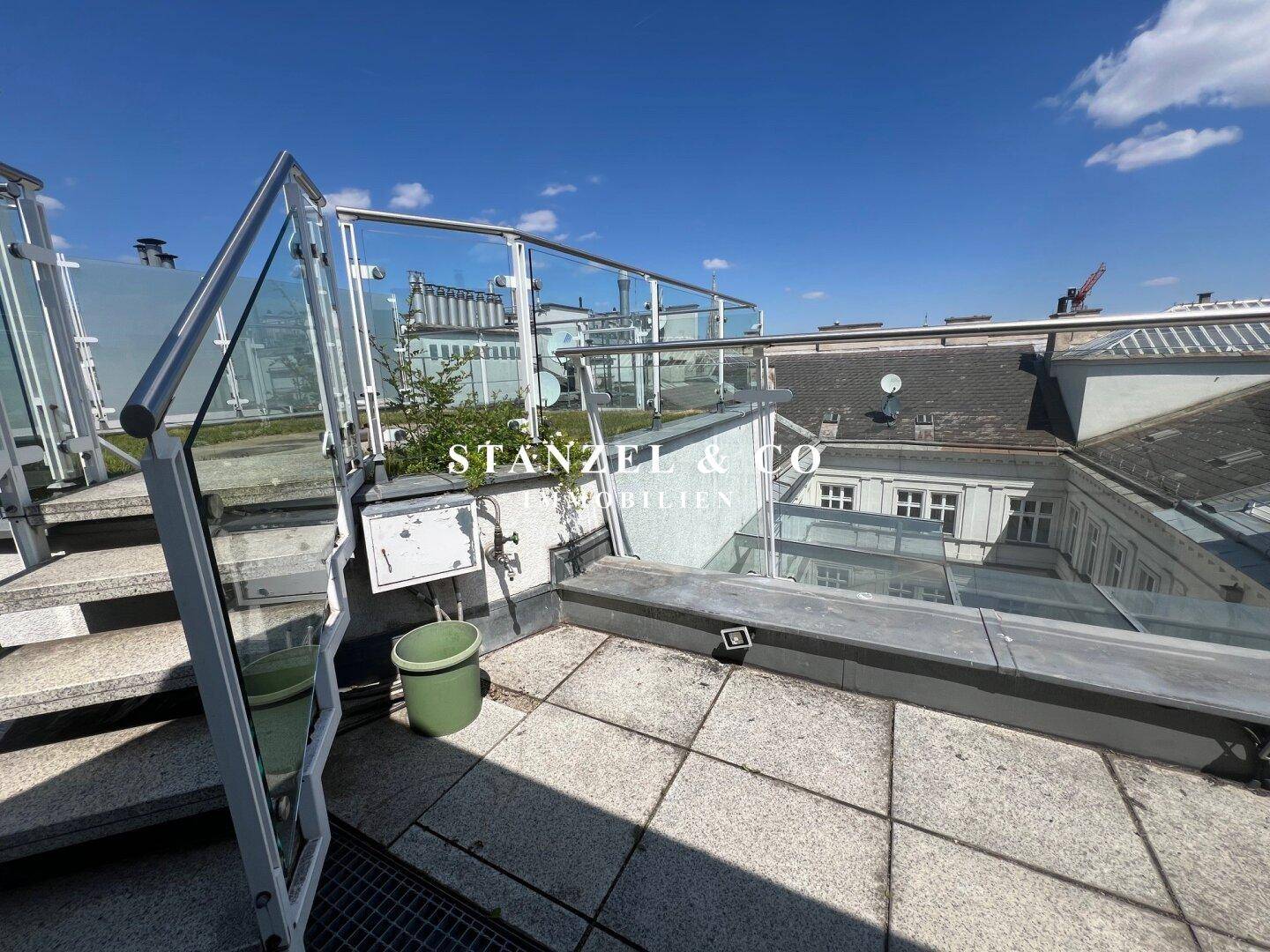Gedeckte Dachterrasse mit Fliesenboden und Treppenzugang, umgeben von Glasbrüstungen.