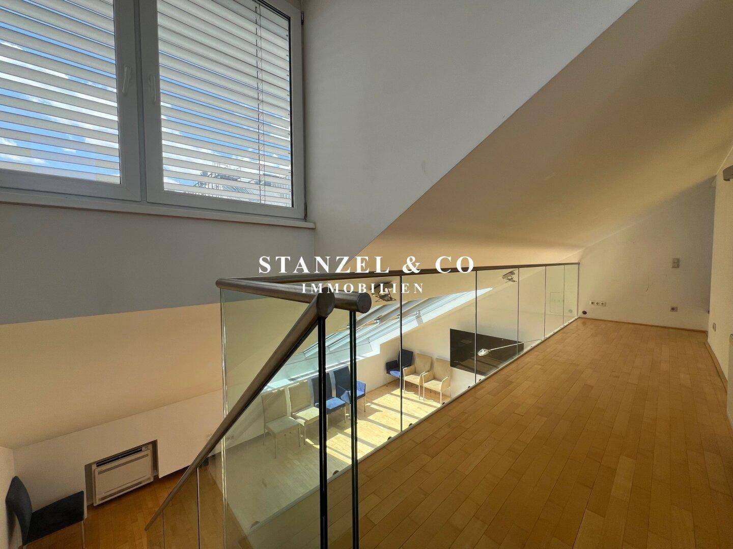 Galeriebereich im Obergeschoss mit Holzboden und Glasgeländer, das den Blick nach unten freigibt.