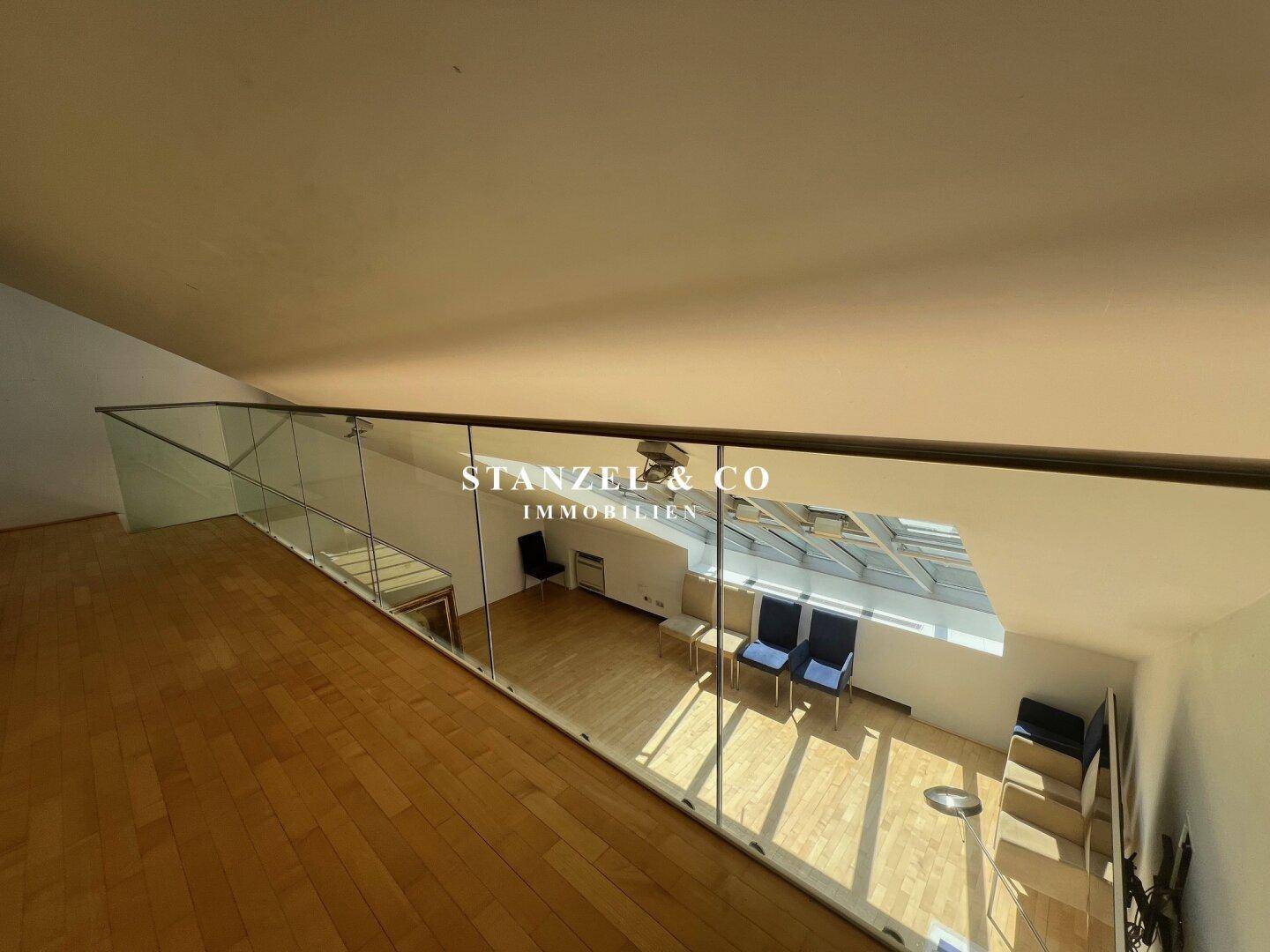 Offener Galeriebereich mit Holzboden und Glasbrüstung, die den Blick auf das Glasdach ermöglicht.