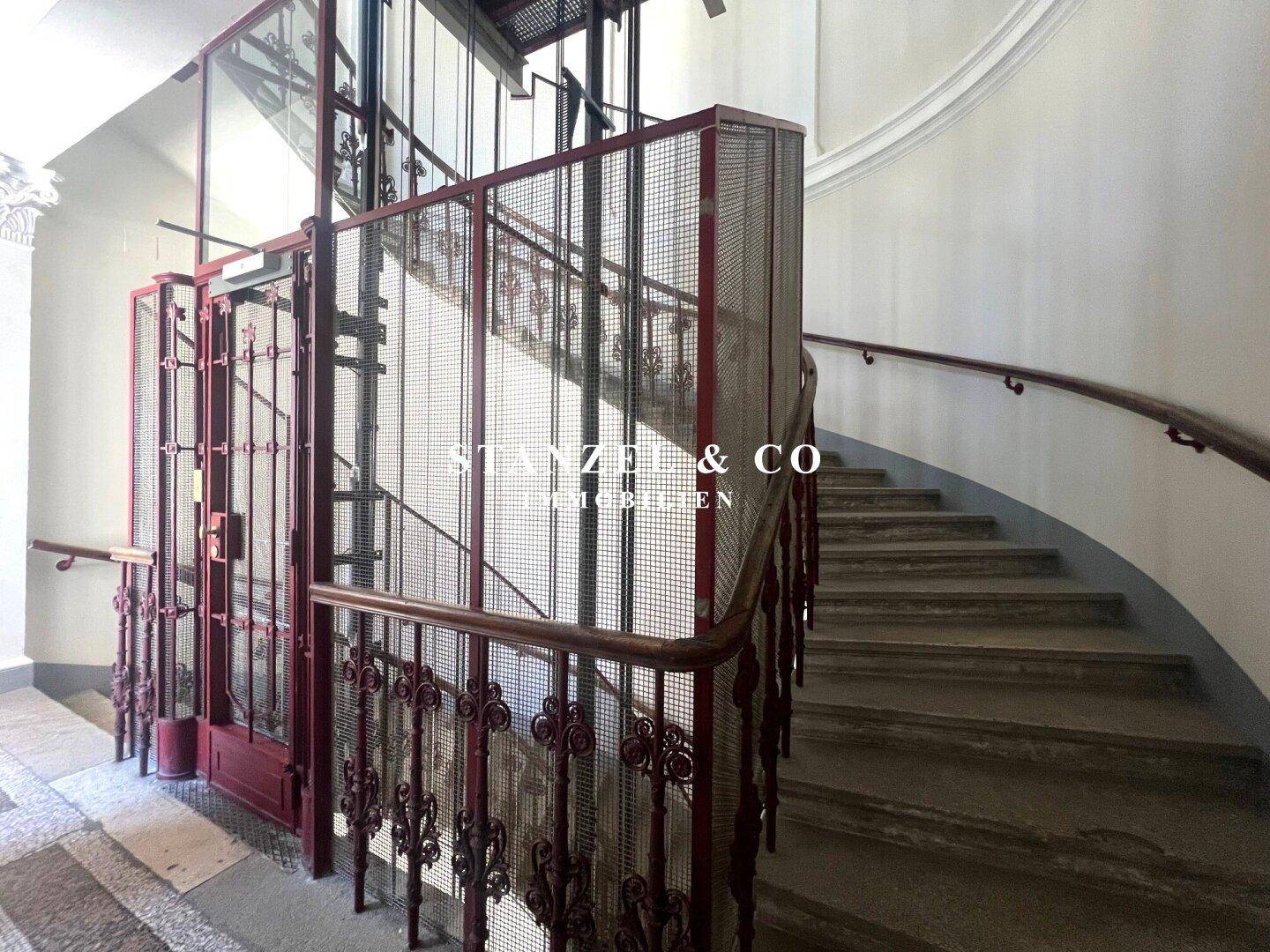 Historischer Aufzug mit kunstvollem Metallgitter neben einer eleganten Steintreppe im Treppenhaus.