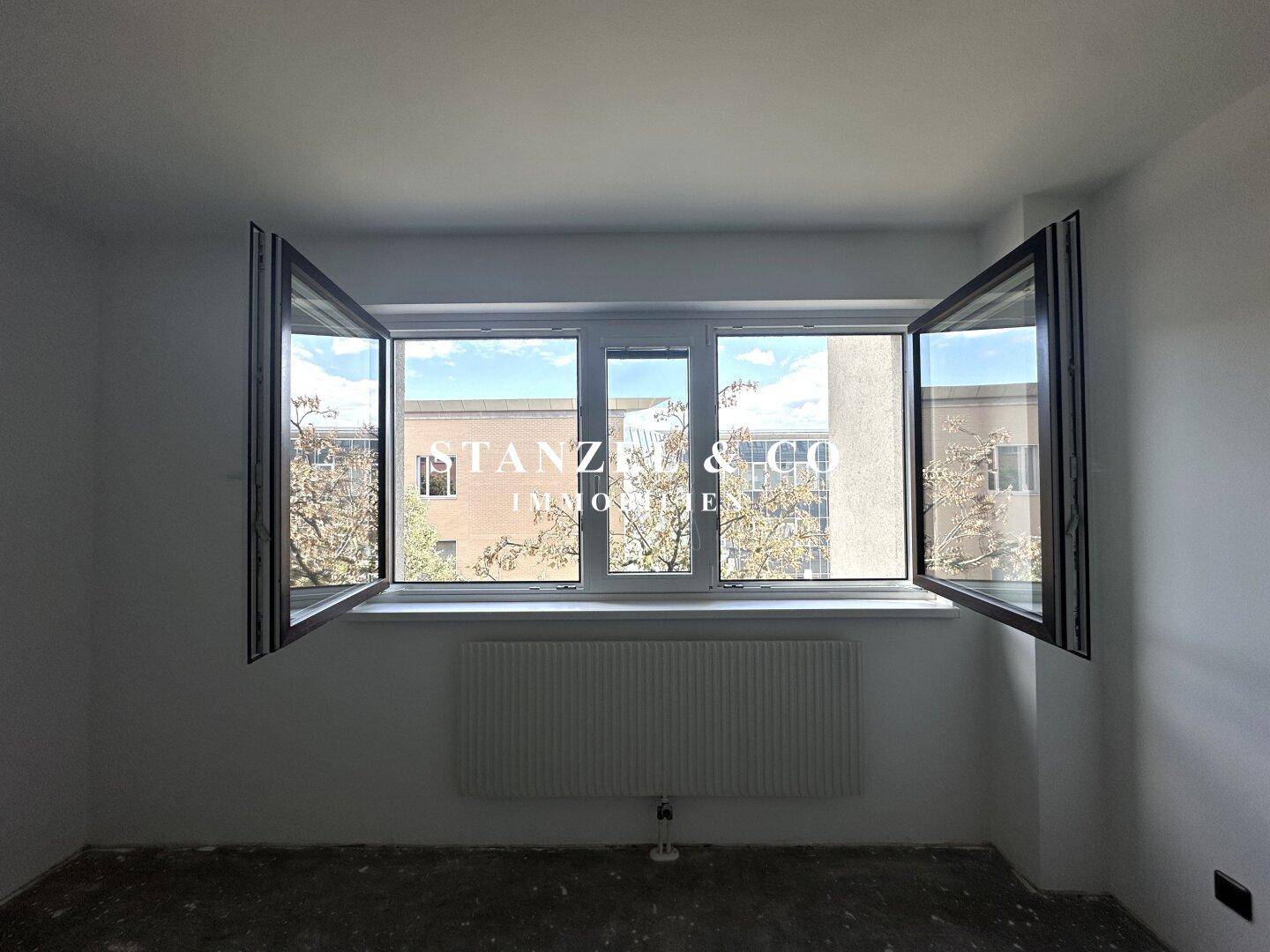 Fensterfront in einem leeren Raum mit Blick auf Bäume und Gebäude im Außenbereich.