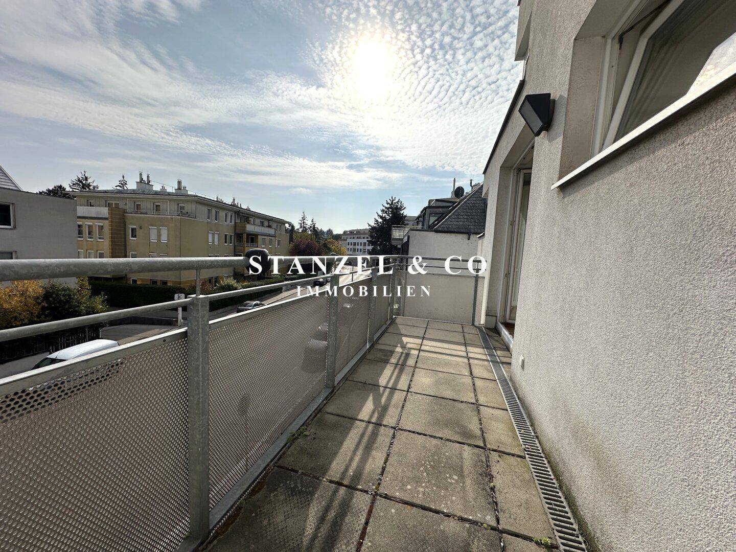 Langer Balkon mit Metallgeländer und Blick auf die umliegenden Gebäude unter sonnigem Himmel.