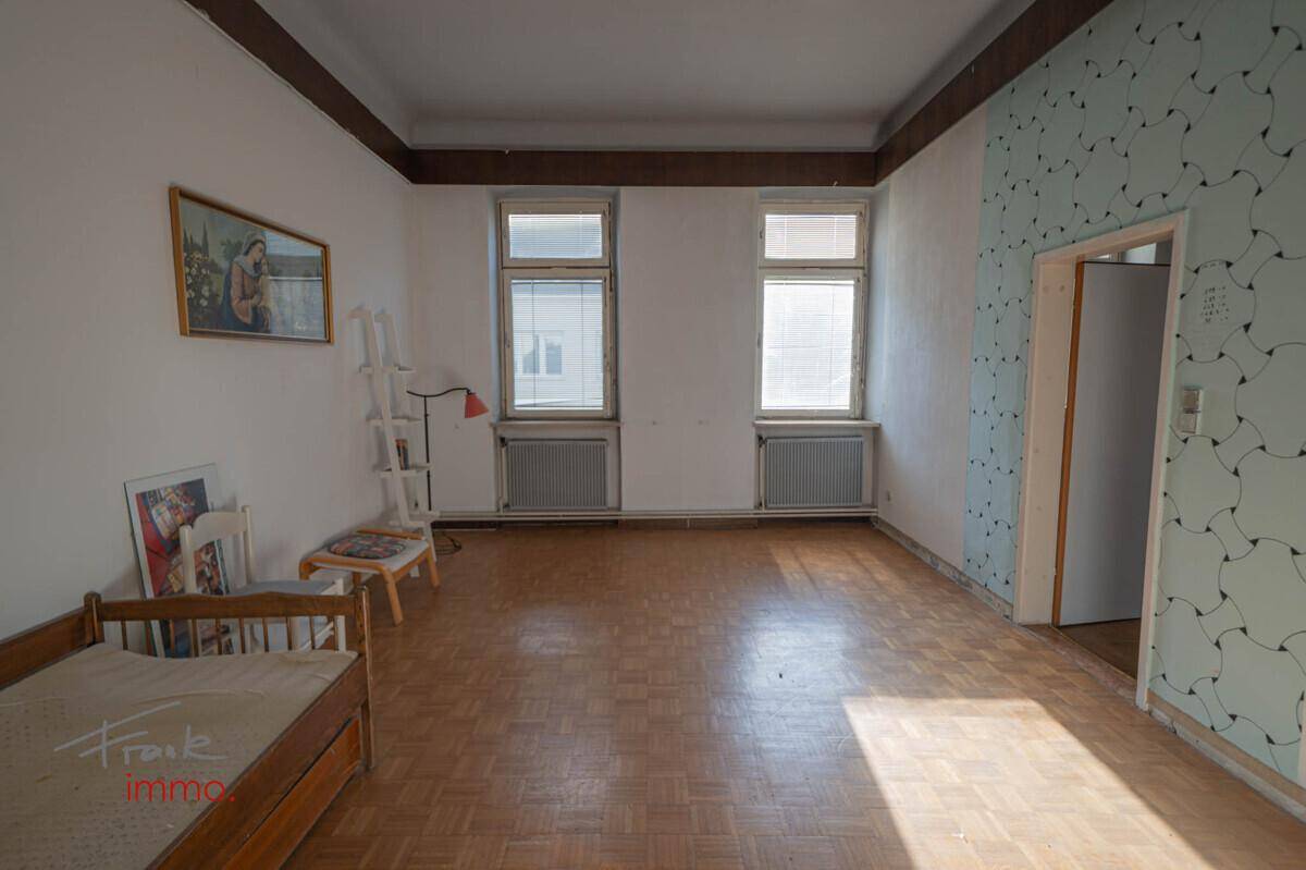 Geräumiger Raum mit Parkettboden, gemusterter Tapete und zwei großen Fenstern.