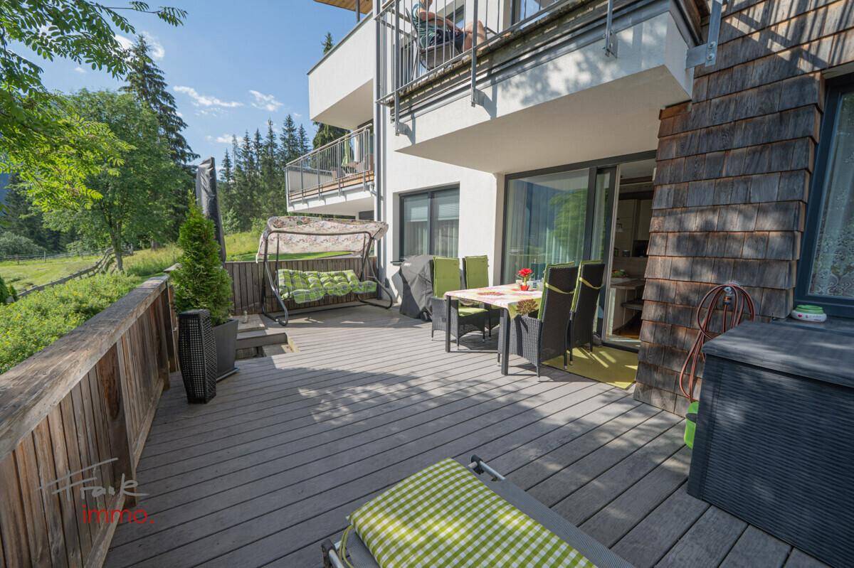Große Holzterrasse mit Schaukel, Esstisch und Liegestuhl, umgeben von Natur.