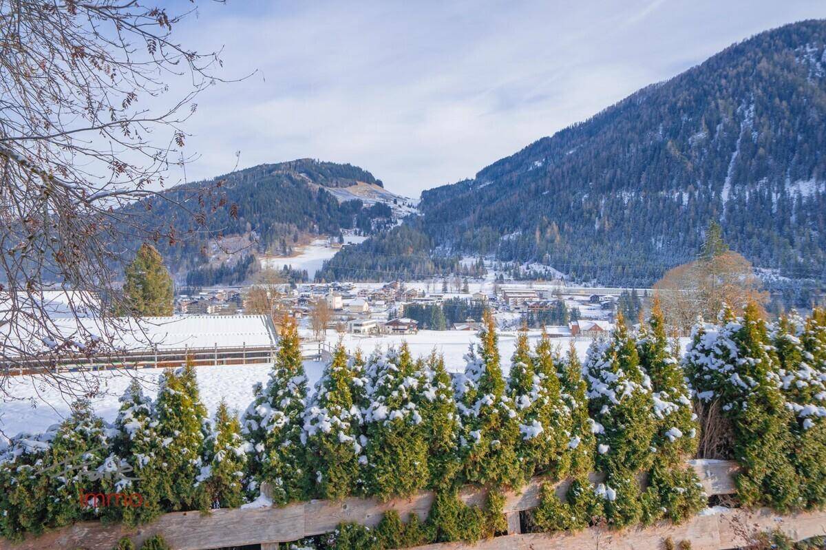 Winterlandschaft mit Blick auf die verschneite Ortschaft und die umliegenden bewaldeten Berge.