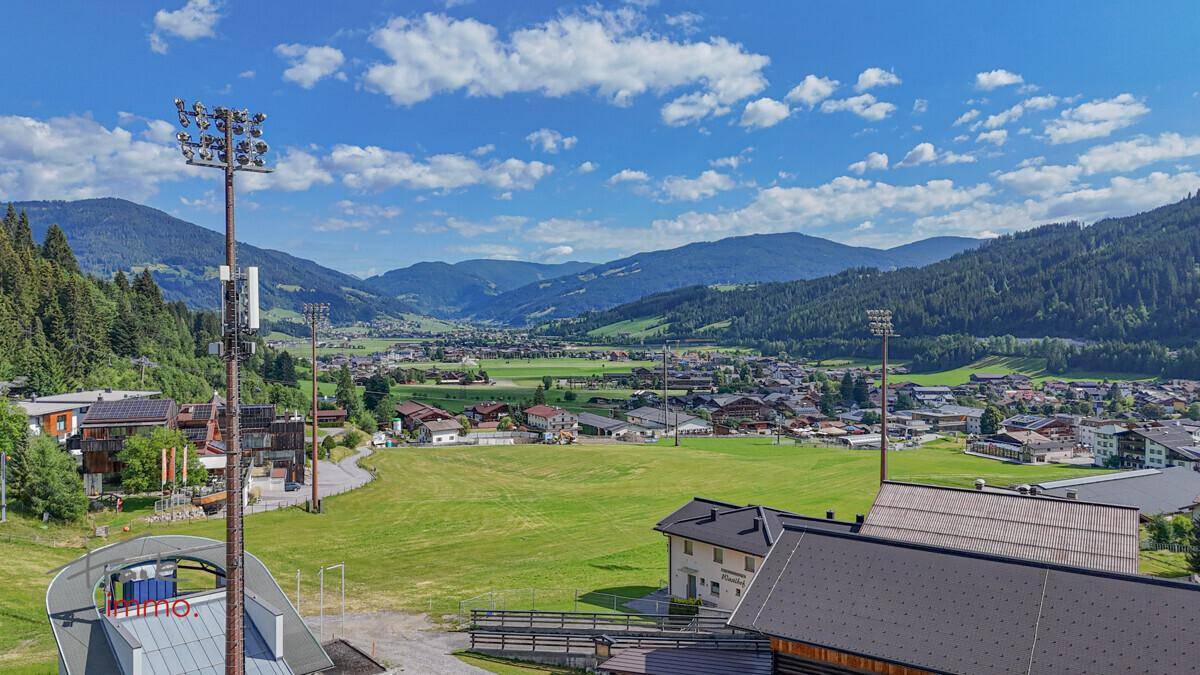 Weitläufiger Blick über das Tal mit grünen Wiesen, Häusern und umliegenden Bergen.