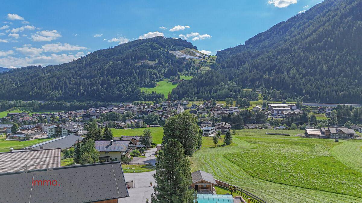 Panoramablick über das grüne Tal mit einer Siedlung und bewaldeten Bergen unter blauem Himmel.