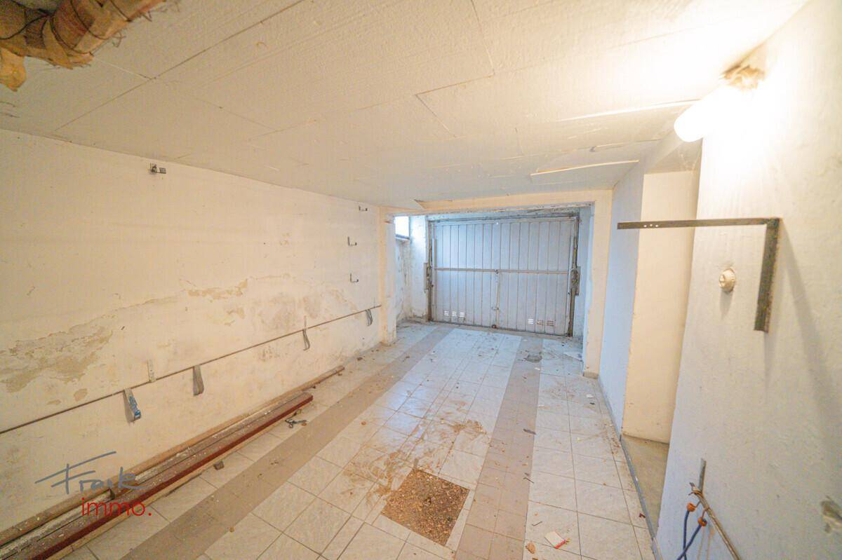 Geräumige Garage oder Lagerraum mit Metalltor, Fliesenboden und sichtbaren Leitungen.