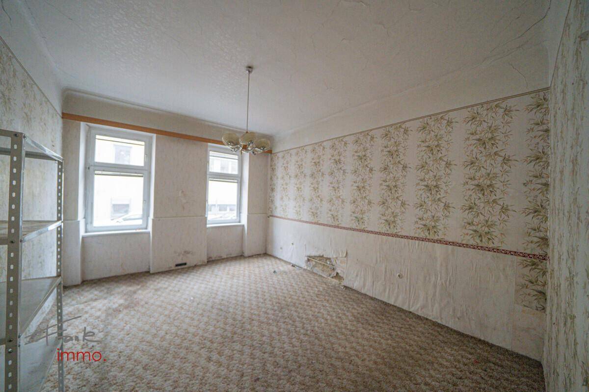 Geräumiger Raum mit alter Tapete, Teppichboden und zwei Fenstern, die Licht hereinlassen.