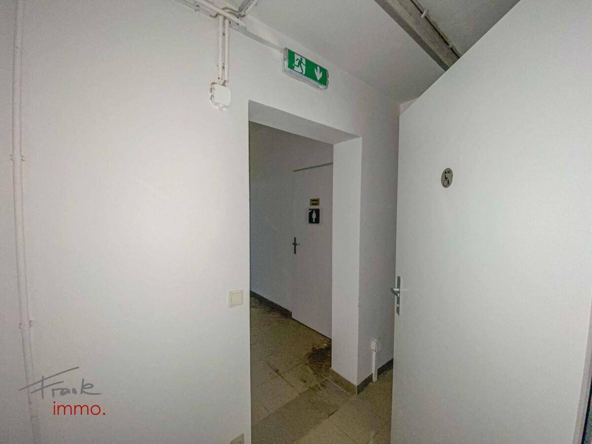 Heller Flur mit weißen Wänden und einem Notausgangsschild, der zu den Toiletten führt.