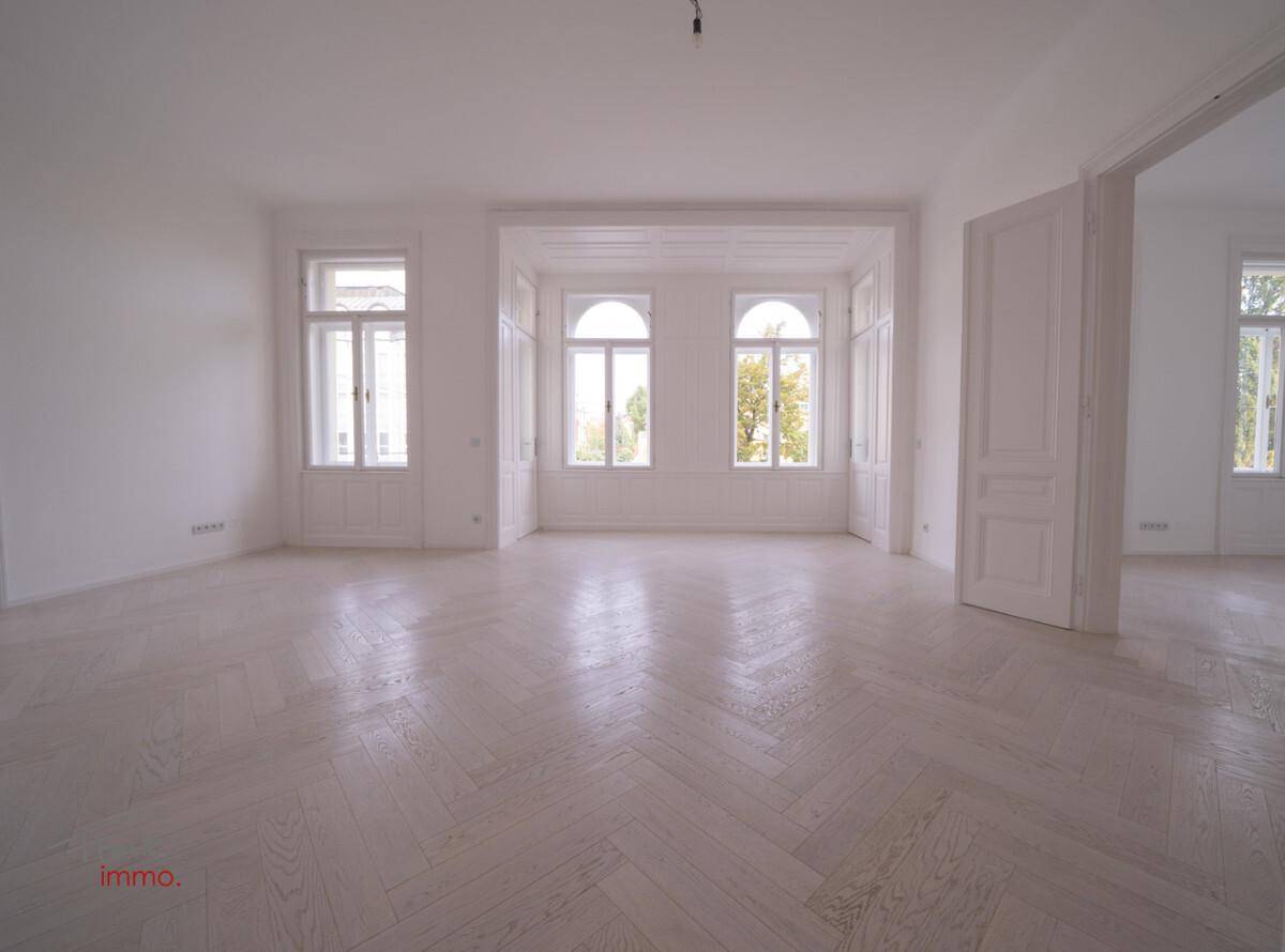 Helles, leeres Zimmer mit Fischgrätparkett, hohen Decken und großen Bogenfenstern, die viel Licht hereinlassen.