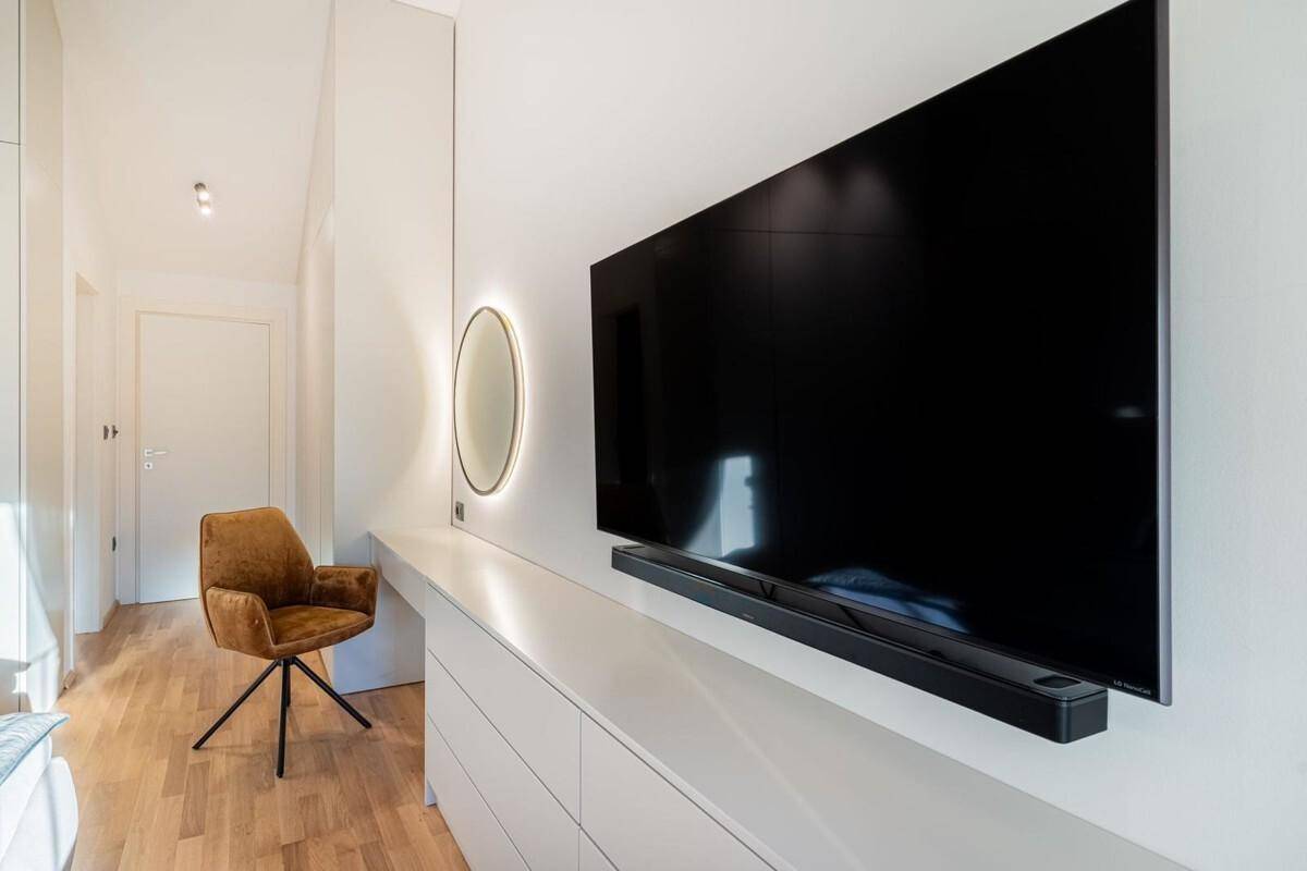 Modernes Schlafzimmer mit Wandfernseher, langer Kommode und beleuchtetem Spiegel.