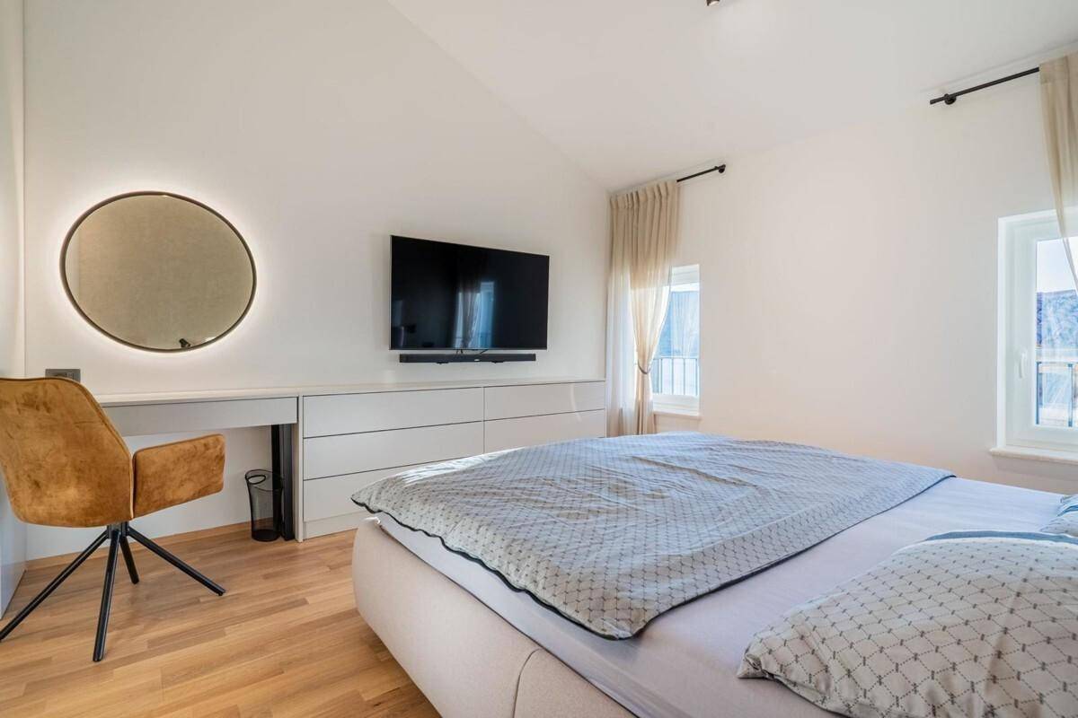 Schlafzimmer mit Holzboden, Wandfernseher und einem beleuchteten runden Spiegel über einer Kommode.