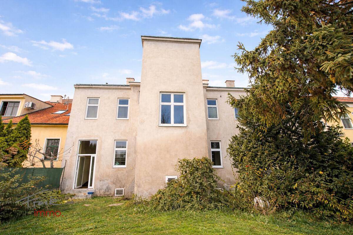 Rückansicht des Gebäudes mit mehreren Fenstern und einem ungepflegten Gartenbereich im Vordergrund.