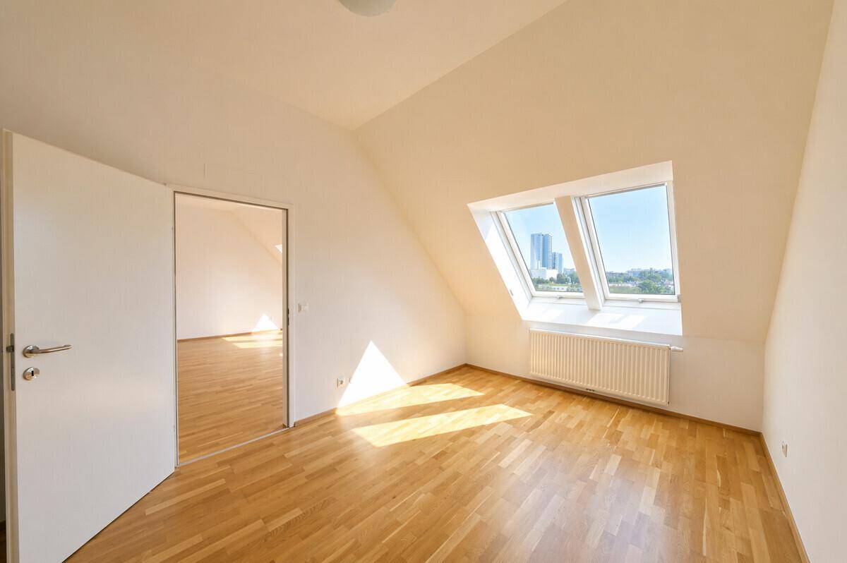 Helles Zimmer im Dachgeschoss mit Parkettboden und Zugang zu einem weiteren Raum, sonnig.