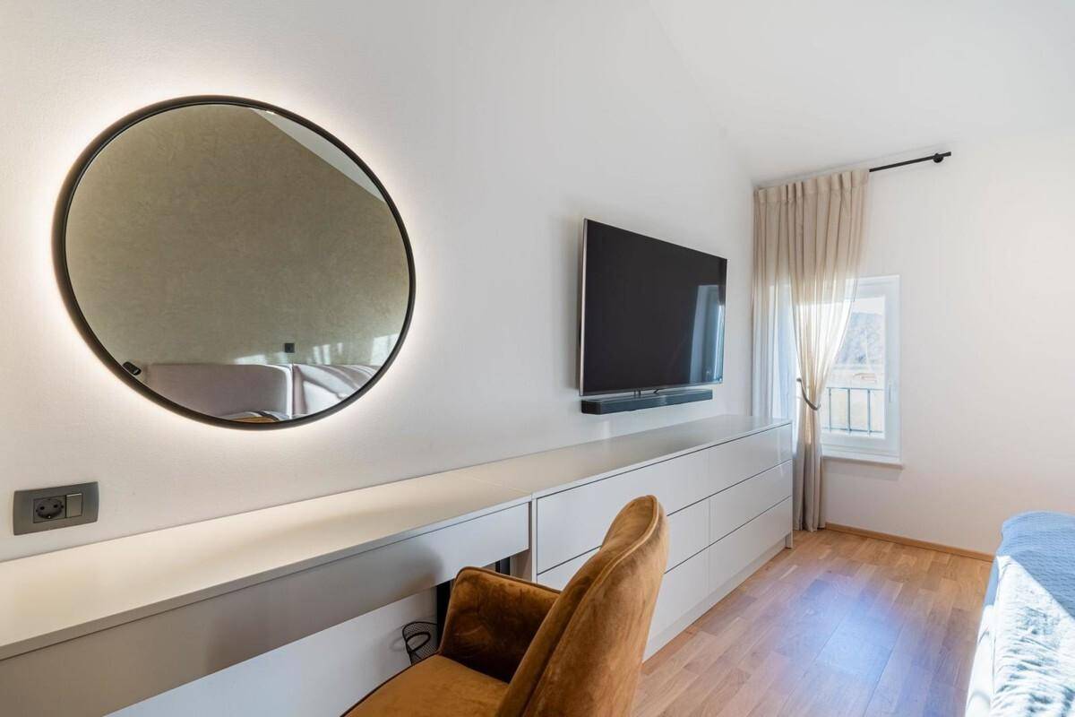 Helles Schlafzimmer mit Holzboden, Wandfernseher und einem beleuchteten runden Spiegel.