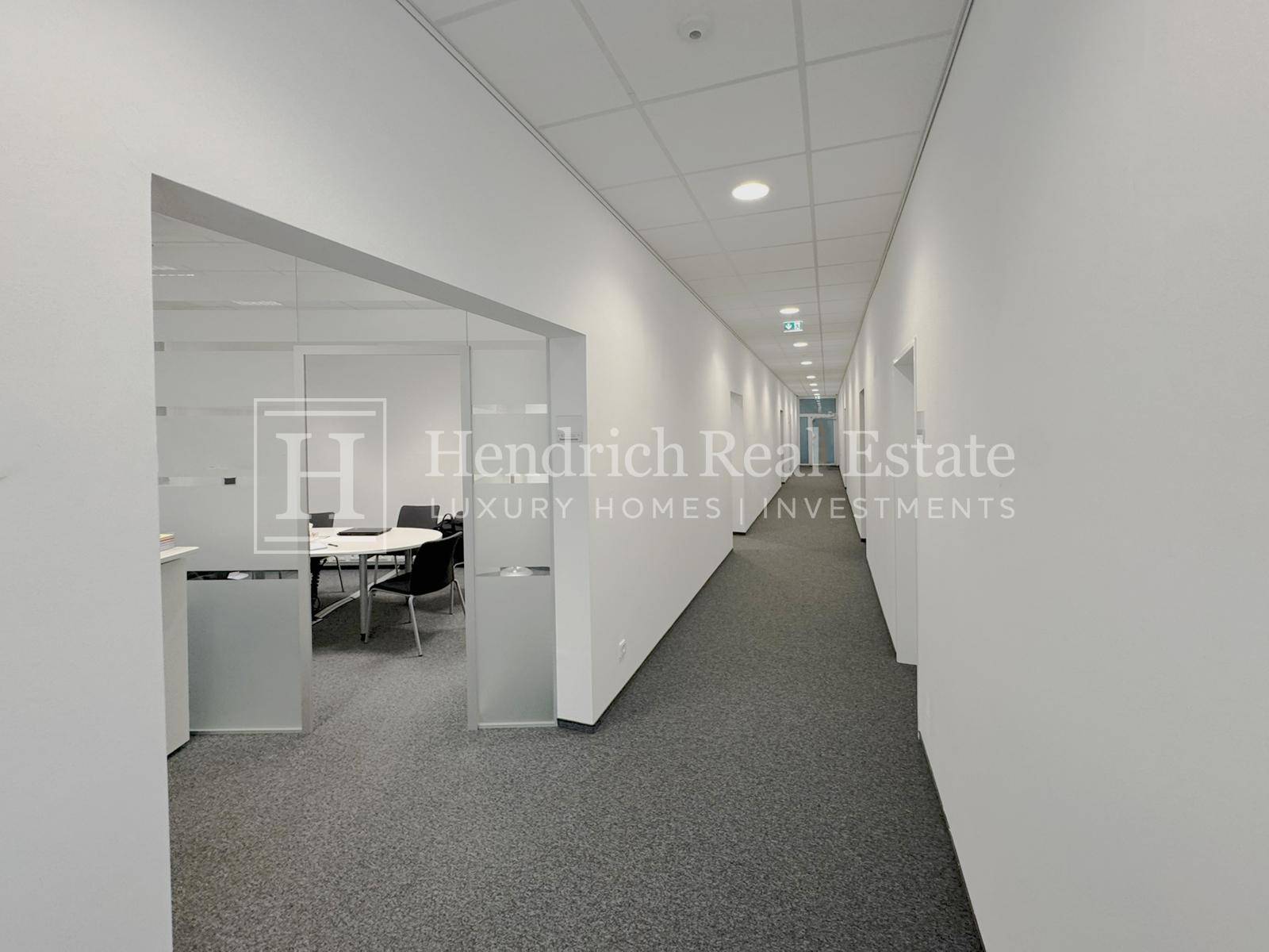 Langer Bürogang mit Teppichboden, weißen Wänden und Blick in einen Besprechungsraum mit Glaswänden.