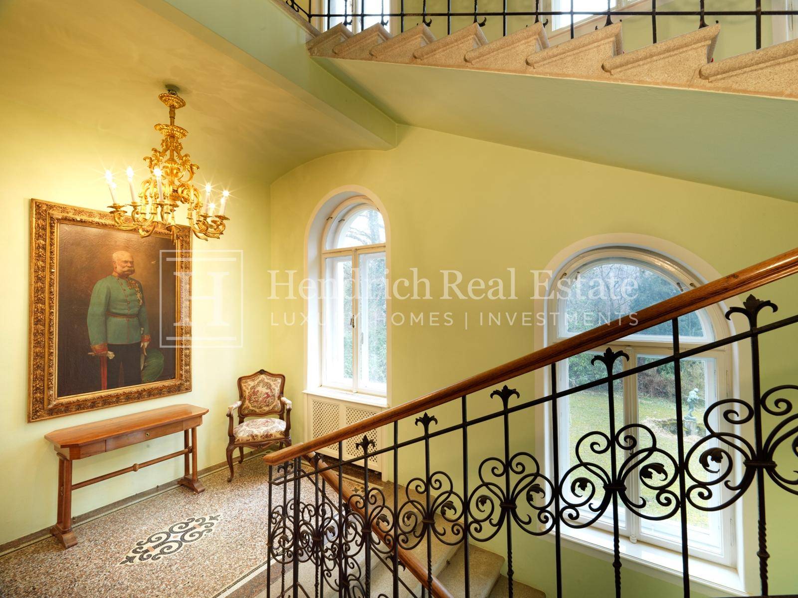 Repräsentatives Treppenhaus mit hohen Bogenfenstern, schmiedeeisernem Geländer und elegantem Kronleuchter.