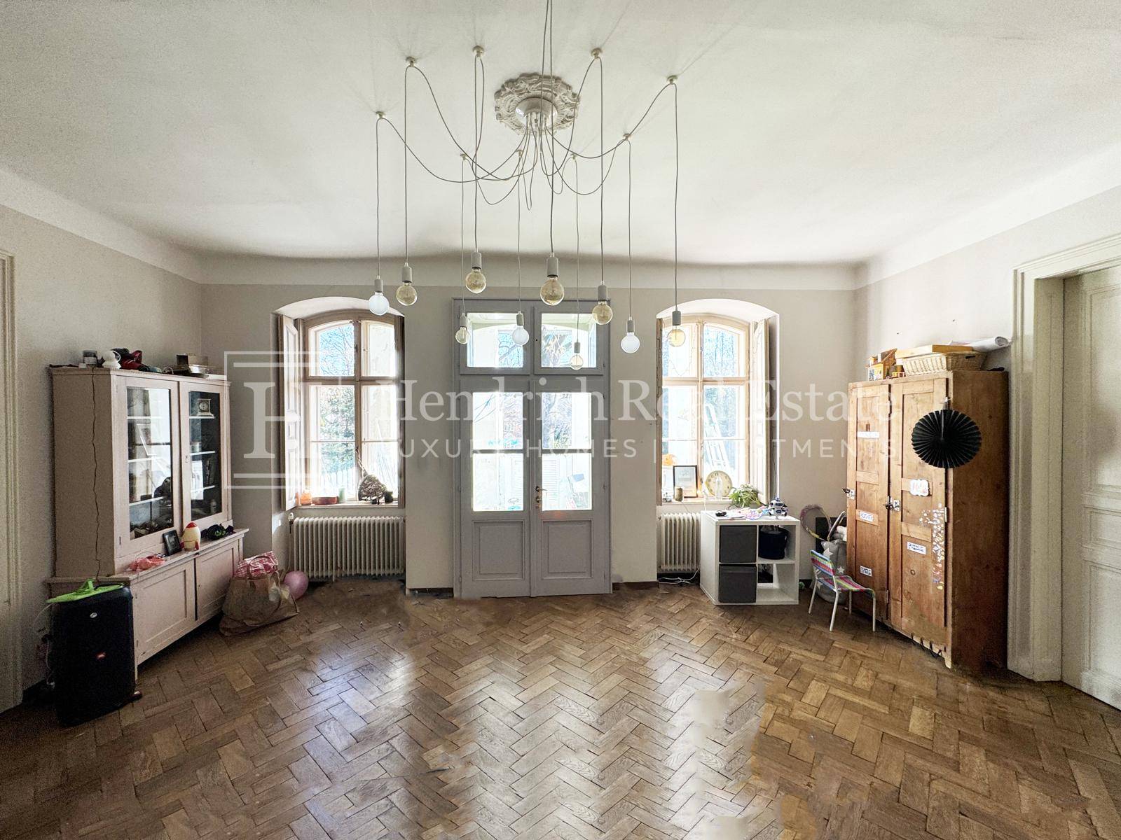 Geräumiges Wohnzimmer mit Fischgrätparkett, hohen Fenstern und dekorativer Deckenrosette.
