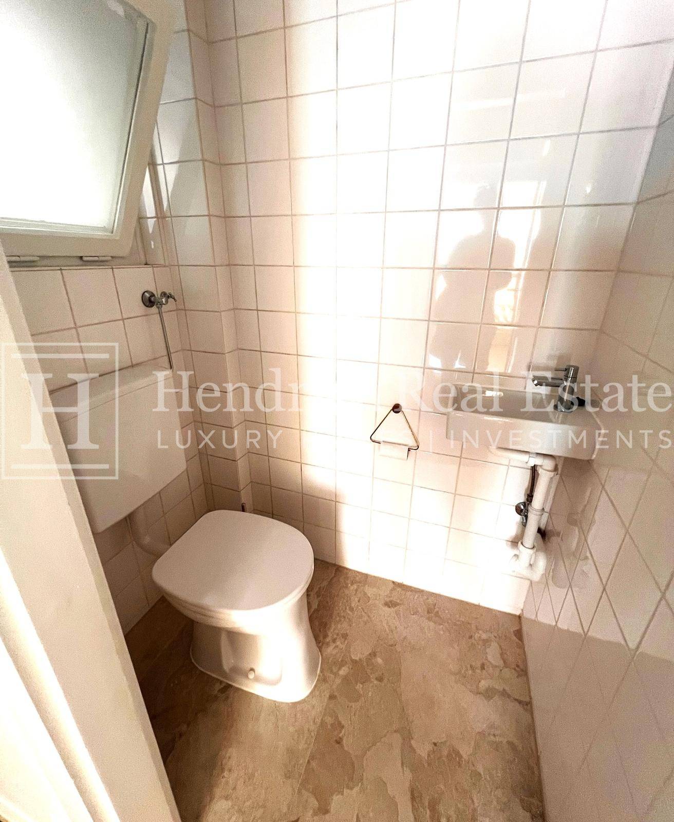 Kleines Gäste-WC mit weißen Fliesen, Toilette, Handwaschbecken und Fenster.