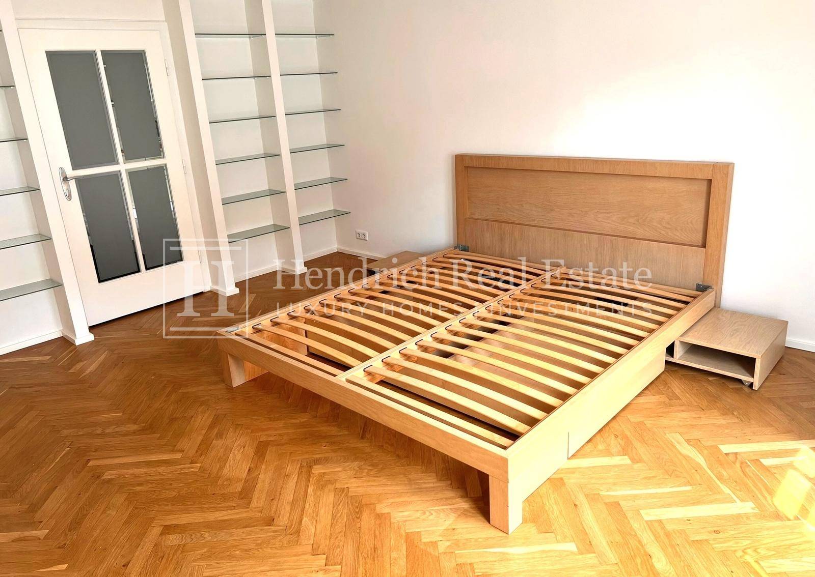 Schlafzimmer mit hellem Holzbettrahmen, integrierten Regalen und klassischem Fischgrätparkett.